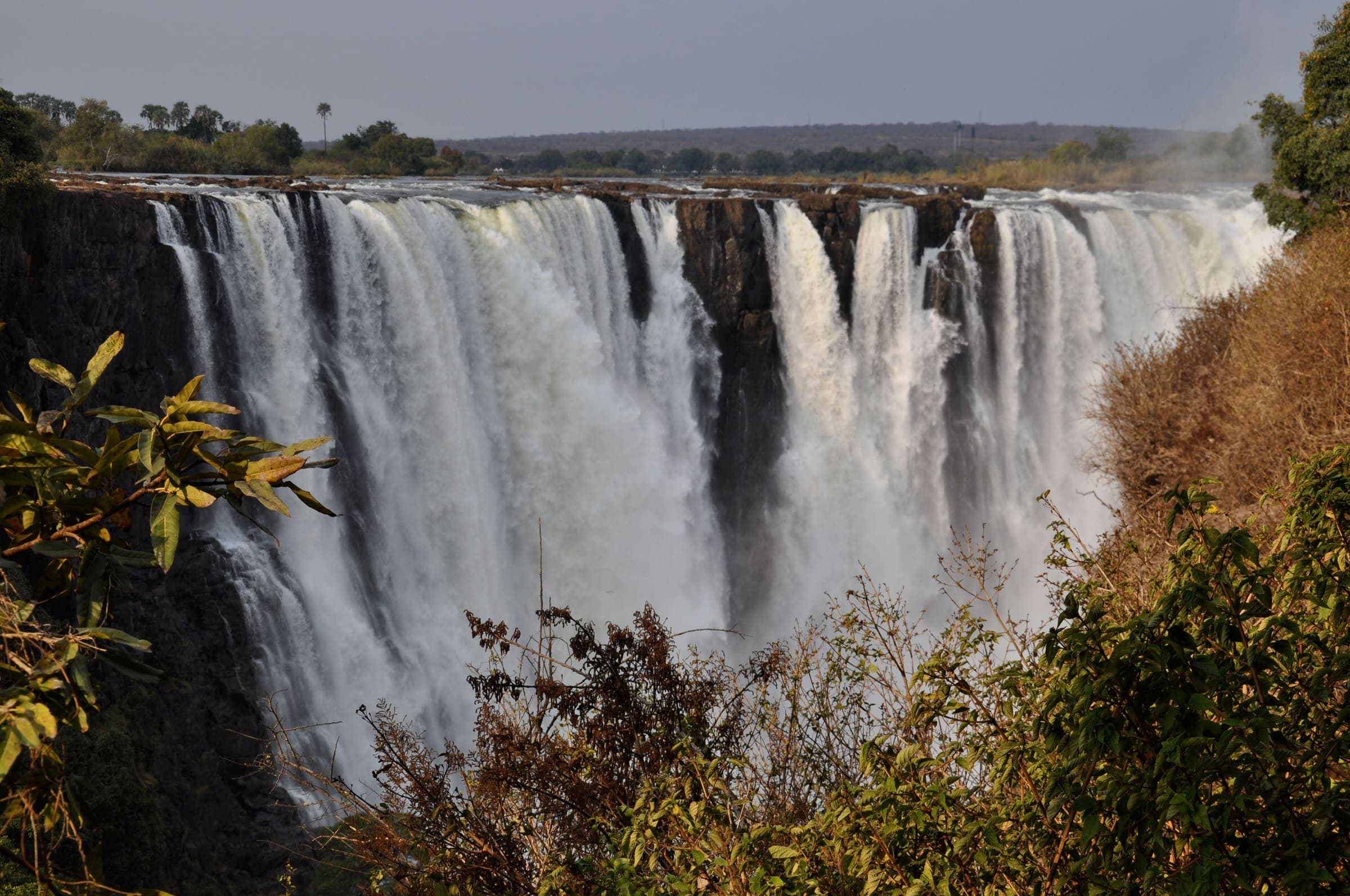 A cortina de água das Cataratas Victoria caindo no desfiladeiro, com a mata seca na borda.