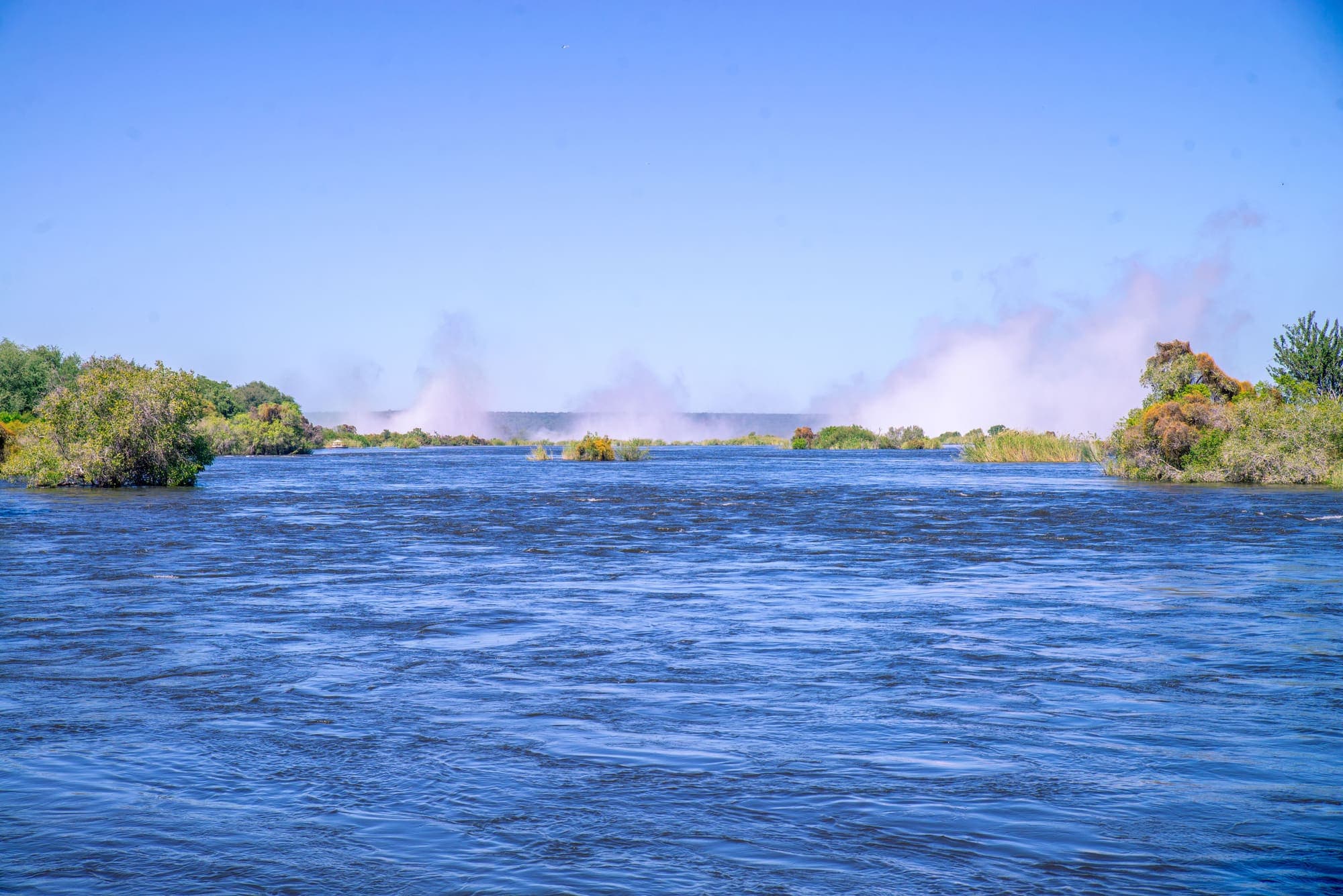O Rio Zambeze largo e azul, com as colunas de névoa das Cataratas Victoria subindo ao fundo.