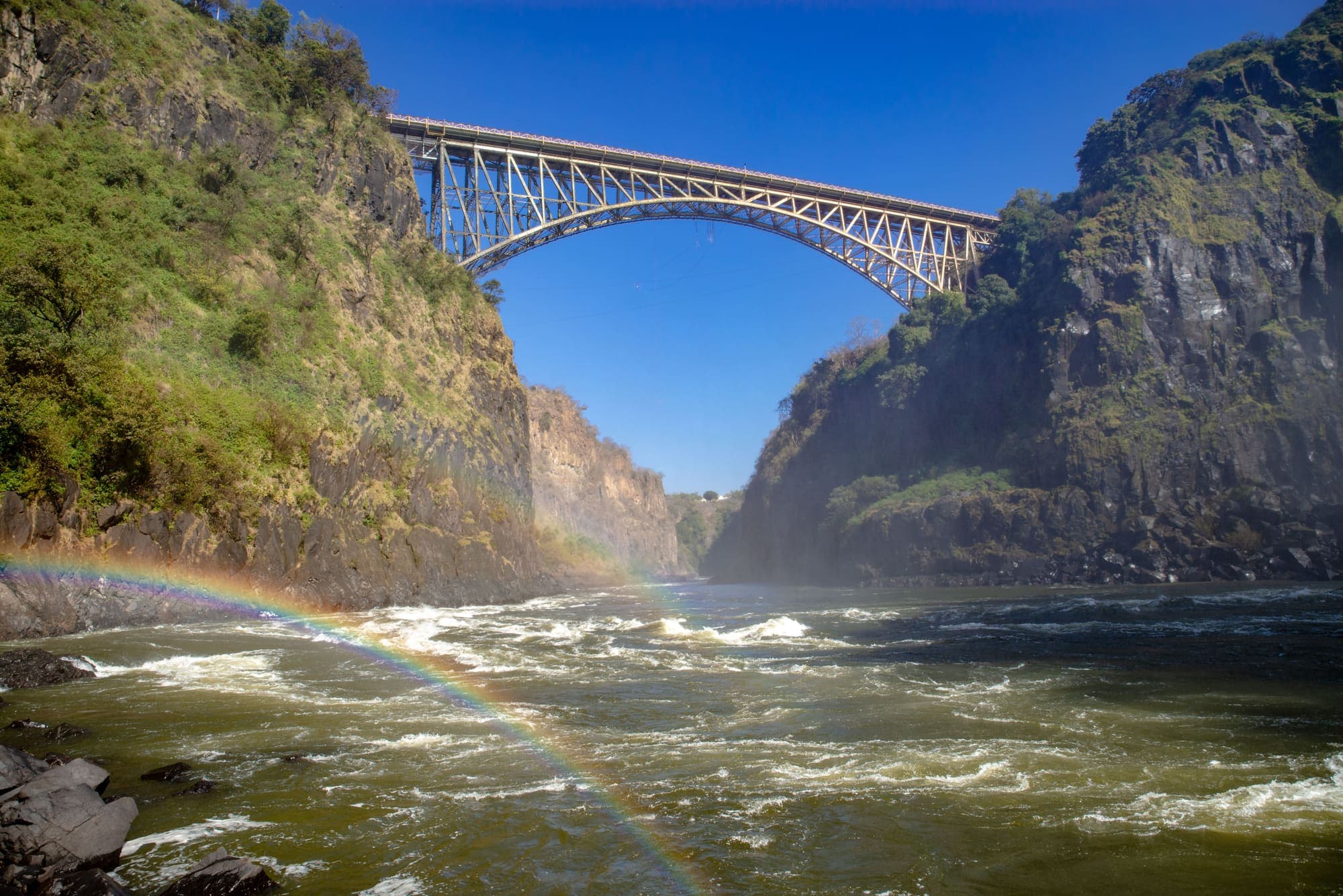 Um arco-íris sobre o Zambeze no fundo do desfiladeiro, com a ponte de Victoria Falls ligando os dois paredões.