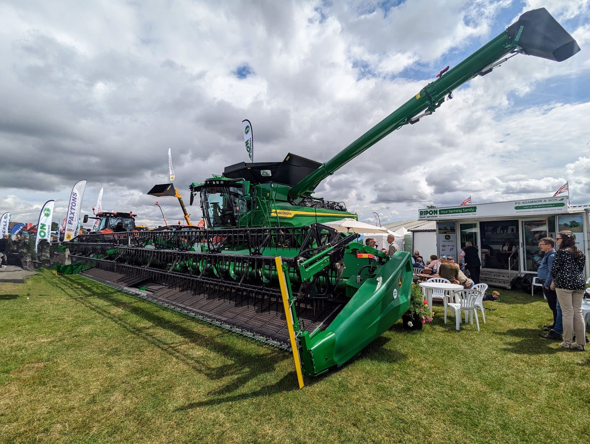 Uma colheitadeira com plataforma larga exposta no gramado de uma feira agrícola ao ar livre, entre tendas e estandes.