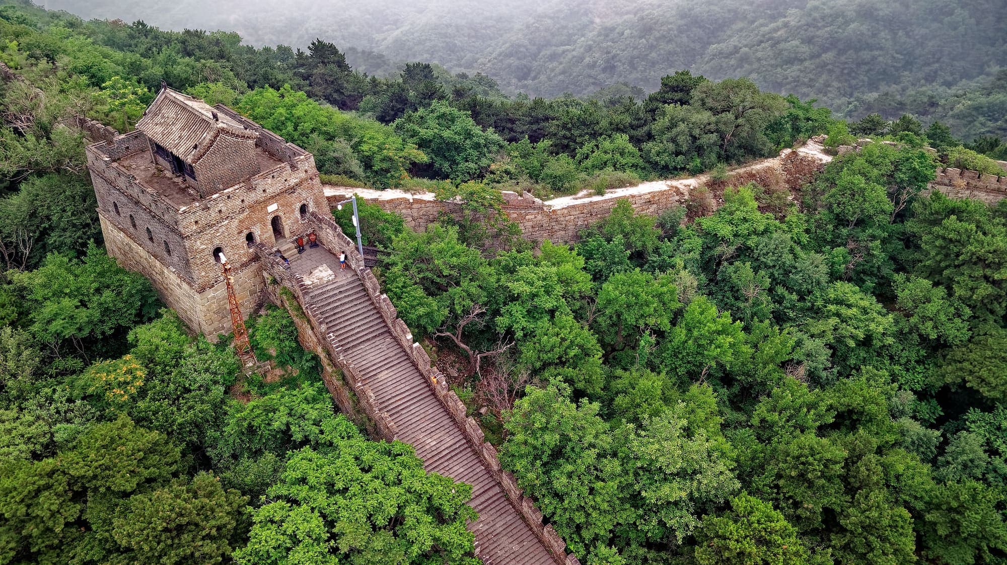 Vista aérea de um trecho restaurado da Grande Muralha em Huairou, ao norte de Pequim, com uma torre de vigia e uma escadaria no meio da mata.