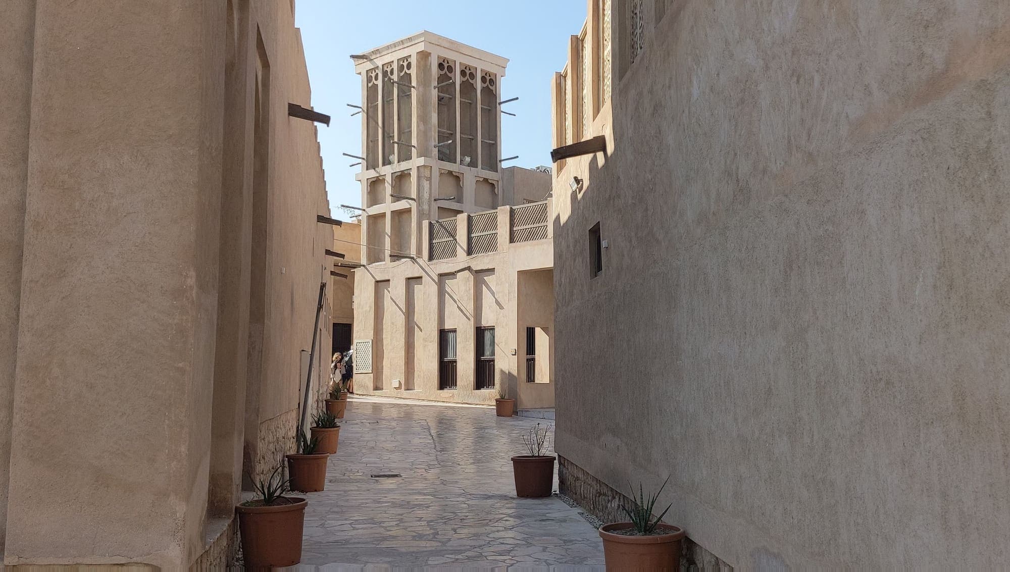 Viela de Al Fahidi, na Dubai antiga, entre casas cor de areia, com uma torre de vento ao fundo.