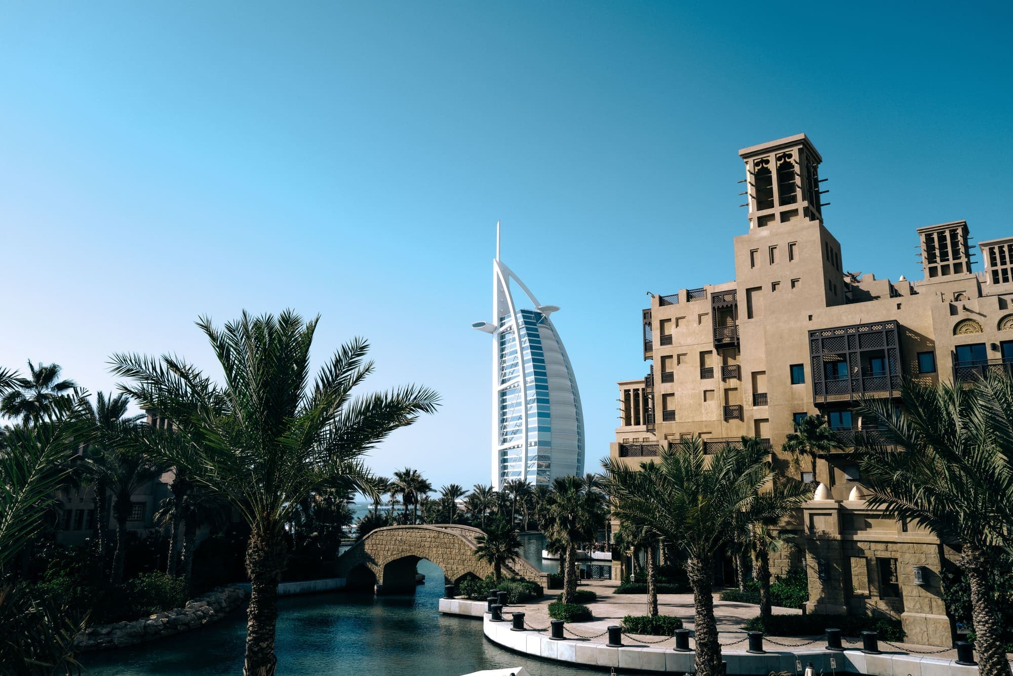 Prédios em estilo árabe com torres de vento e palmeiras na beira de um canal em Jumeirah, com o Burj Al Arab ao fundo.