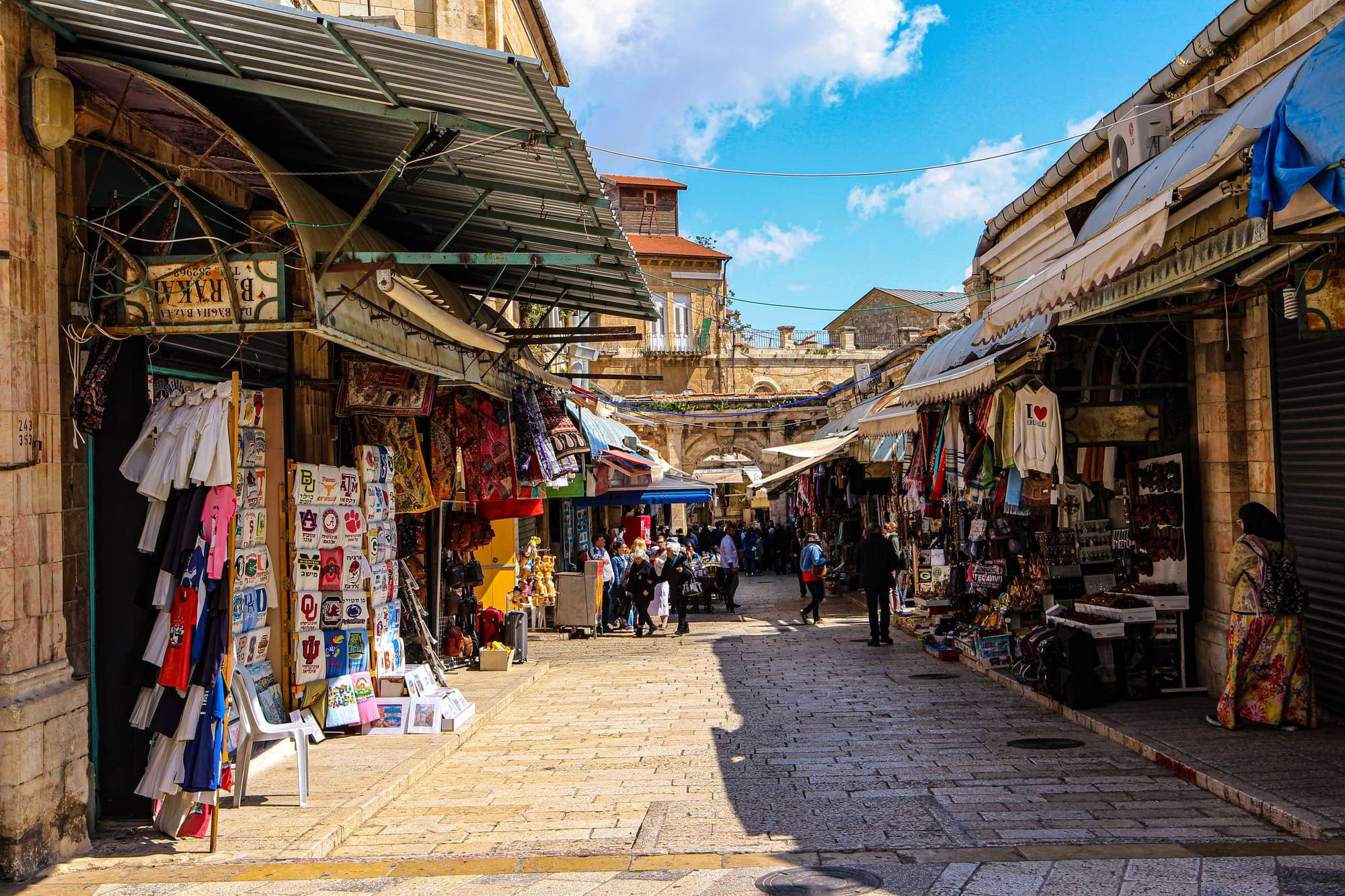 Uma rua de comércio da Cidade Velha de Jerusalém, com lojas de lembranças e toldos dos dois lados.
