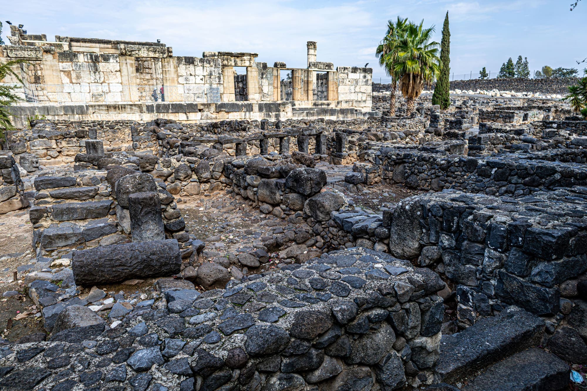 As ruínas de Cafarnaum: casas de pedra basáltica escura em primeiro plano e a sinagoga de calcário branco ao fundo.