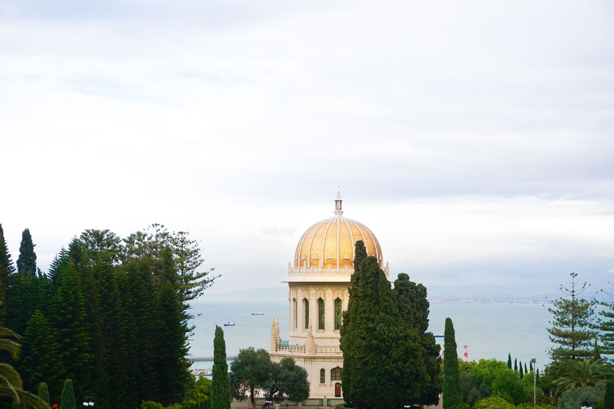 O Santuário do Báb, de cúpula dourada, entre ciprestes nos Jardins Bahá'í, em Haifa, com o mar ao fundo.