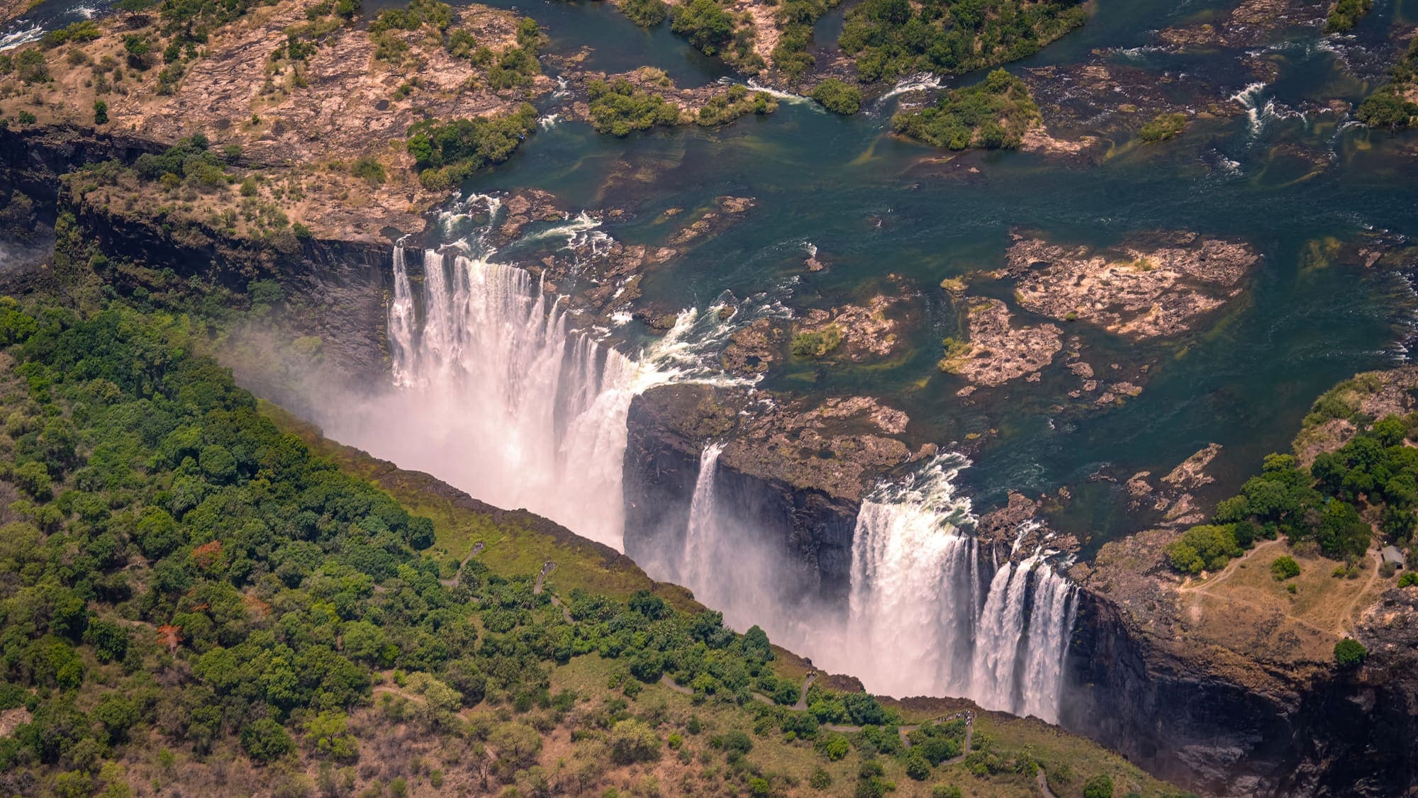 Vista aérea das Cataratas Victoria, com o Rio Zambeze despencando no desfiladeiro cercado de mata.