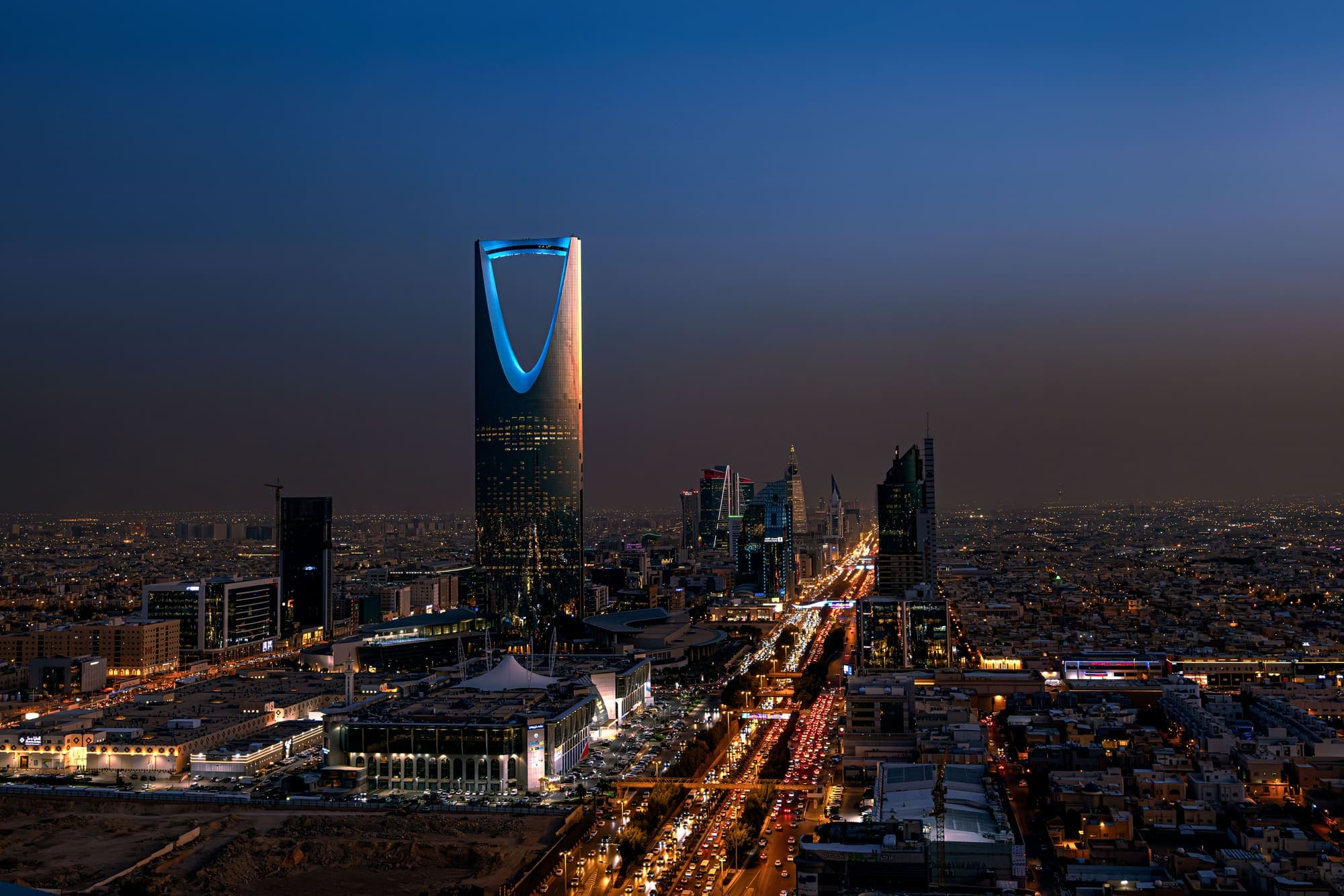 Vista aérea de Riade ao anoitecer, com o Kingdom Centre iluminado e os prédios do centro financeiro ao fundo.