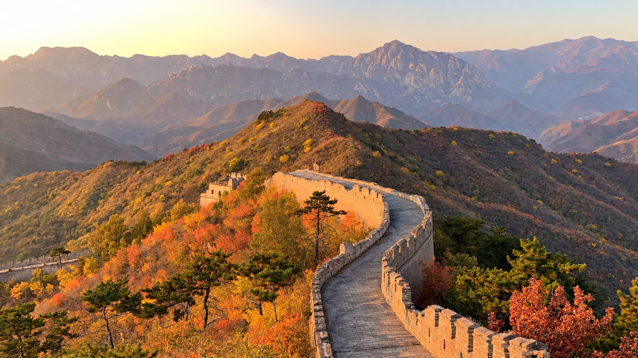 A Grande Muralha da China serpenteando pela crista de um morro de árvores em tons de laranja e vermelho, com montanhas ao fundo.