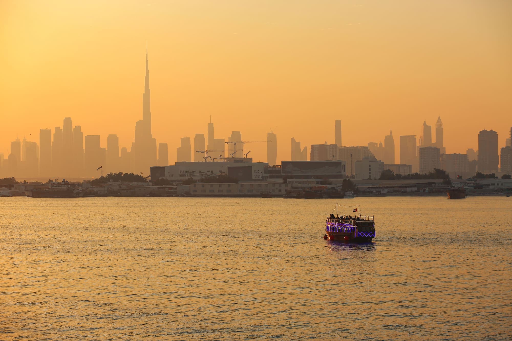 O horizonte de Dubai, com o Burj Khalifa, sob o céu dourado do entardecer, e um barco tradicional iluminado navegando em primeiro plano.
