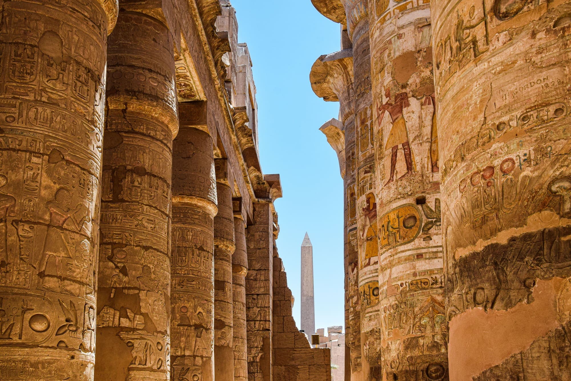 As colunas da Sala Hipostila do Templo de Karnak, em Luxor, cobertas de hieróglifos, com um obelisco ao fundo.