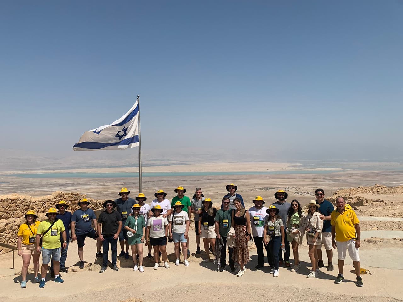 Grupo Adama com chapéus amarelos no alto da Fortaleza de Massada, junto à bandeira de Israel, com o Mar Morto ao fundo.