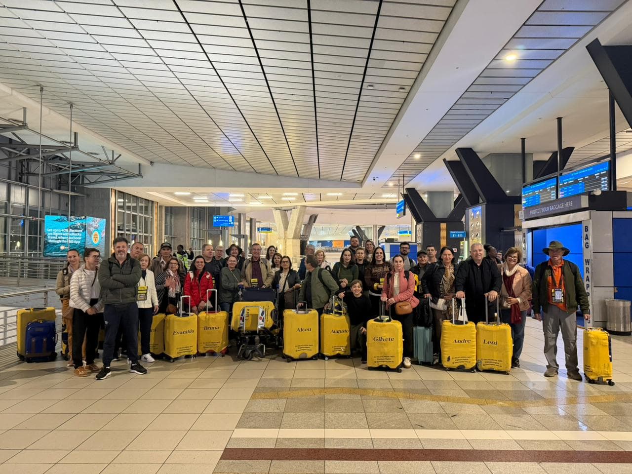 Grupo da Leão Viagens no saguão de um aeroporto, com as malas em capas amarelas da Leão marcadas com o nome de cada viajante.